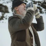 Senner in Loden-Wollmantel mit Nordisches-Muster-Handschuhen und Strickmütze vor verschneiter Ehrwalder Winterlandschaft, eriro Alpine Hide