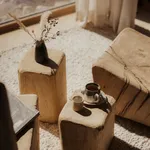 Massivholz-Beistelltische aus handgeschnitzten Baumstämmen mit Keramiktasse und getrockneten Gräsern im eriro Alpine Hide – warme alpine Wohnkultur