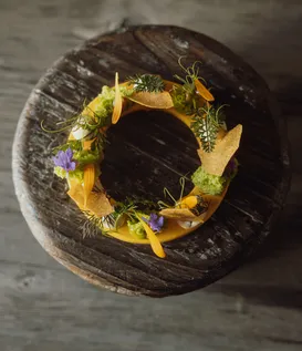 Kunstvoll kreisförmig angerichtetes Gericht mit essbaren Blüten und Kräutern auf verwittertem Holzblock – alpine eriro Fine-Dining-Kulinarik