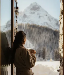 Woman in bathrobe with cup at window of eriro Alpine Hide gazing at snow-covered winter landscape with mountain panorama near Zugspitze