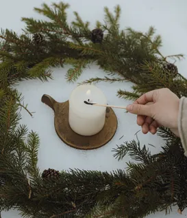 Hand lighting white pillar candle on brass dish, framed by spruce branch wreath with cones on fresh snow – quiet eriro winter ceremony