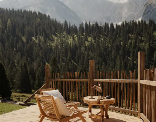 Sonnendurchflutete Holzterrasse mit Rattanstuhl und Bergpanorama – stille Morgenmomente inmitten der Alpenwälder im eriro Alpine Hide