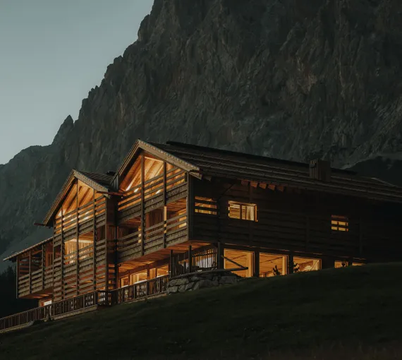 eriro Alpine Hide bei Dämmerung mit goldbeleuchteten Lärchenloggien und horizontalen Holzlamellen vor schattigem Wettersteinmassiv auf Almhang