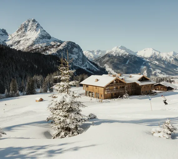 eriro Alpine Hide – Winteridylle mit Skifahrer vor dem Luxuschalet inmitten tiefverschneiter Alpenlandschaft mit Bergpanorama und gezuckerten Tannen