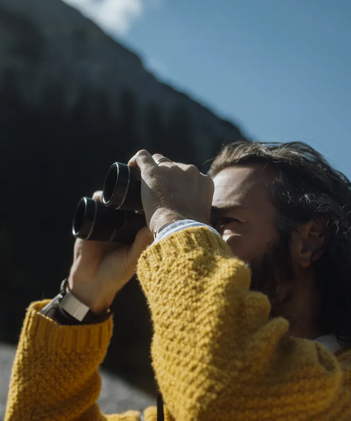 Gast im senfgelben Strickpullover blickt durch ein Fernglas in die Alpenwelt – achtsame Naturbeobachtung im eriro Alpine Hide
